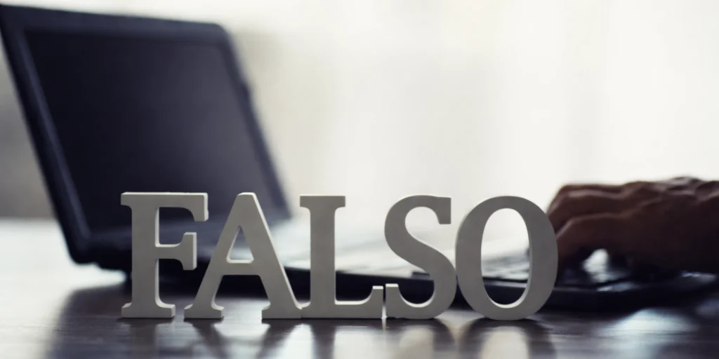 La palabra «FALSO» formada por letras blancas sobre un escritorio oscuro, con un portátil abierto y una persona escribiendo desenfocados en el fondo.